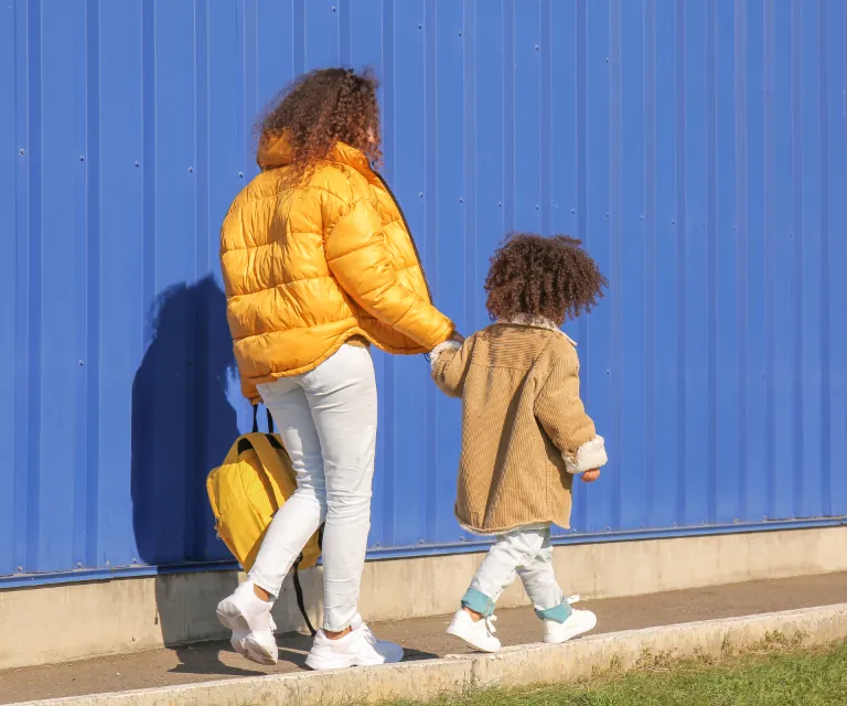 Mutter und Kind gehen Hand in Hand an einer blauen Wand entlang, von hinten